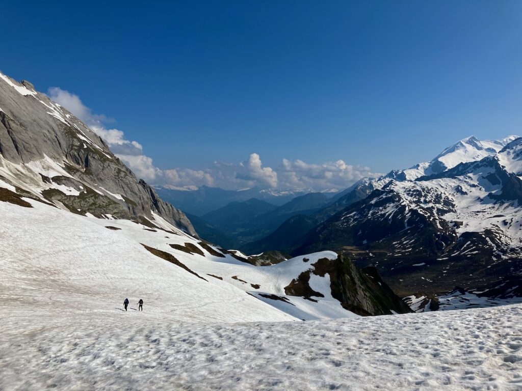 A landscape with a clear blue sky and a snow-covered mountain slope, with two people seen lower down the slope hiking up the slope, their small size in the picture emphasizing the grandness of the view