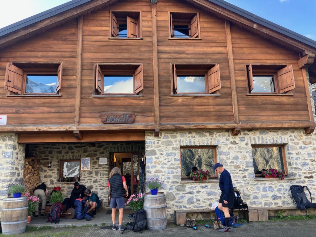 A large mountain hut built up of a stone ground floor and a wooden top floor with four windows. A sign says "Rifugio Bonatti". Hikers in front of the building are preparing for the day, packing their backpacks.